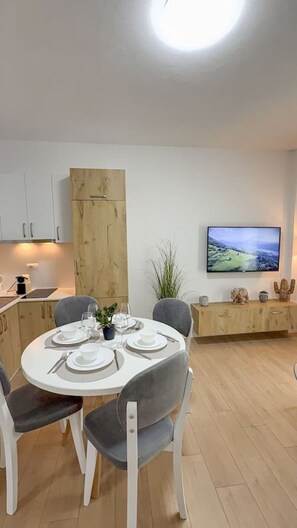 A modern kitchen with white cabinets, a wooden cabinet, and a dining area with a round table set for four. - Tiara Beach Apartment (Durrës)