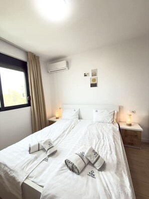 A double bed with white linens, two bedside tables with lamps, an air conditioning unit, and a window with curtains. - Tiara Beach Apartment (Durrës)