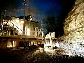 A wooden cabin with a staircase leading to a deck, surrounded by trees and a rock formation. - Spacious Tree House near Starved Rock and Buffalo Rock State Parks, Perfect for a Family Glamping Getaway (Ottawa)