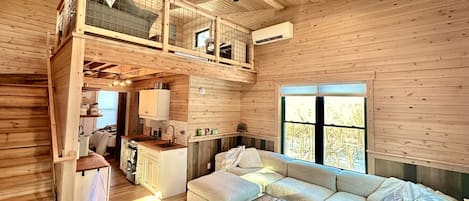 A wooden cabin interior with a loft bedroom, a kitchen, a living area with a sofa and coffee table, and a ceiling fan.