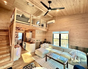 A wooden cabin interior with a loft bedroom, a kitchen, a living area with a sofa and coffee table, and a ceiling fan. - Spacious Tree House near Starved Rock and Buffalo Rock State Parks, Perfect for a Family Glamping Getaway (Ottawa)