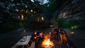 A rustic outdoor setting with a campfire, a picnic table, and a wooden deck overlooking a forested area.