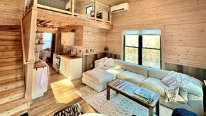 A wooden cabin interior with a loft bedroom, a kitchen, a living area with a sofa and coffee table, and a ceiling fan.