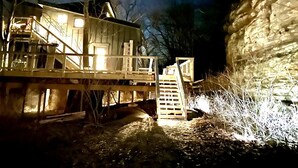 A wooden cabin with a staircase leading to a deck, surrounded by trees and a rock formation.