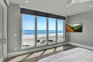 Ein Schlafzimmer mit großem Fenster mit Blick auf einen Strand, einem Wand-Fernseher und einem gemusterten Teppich.