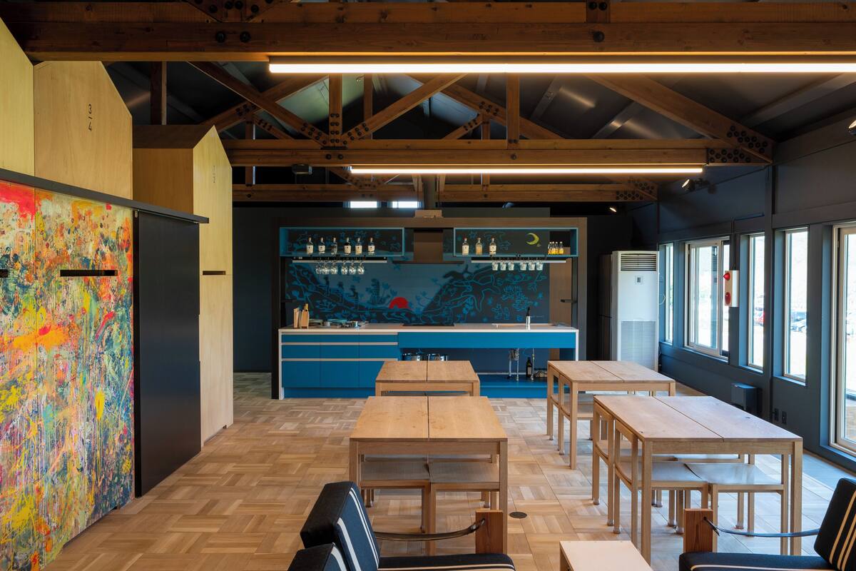 A modern interior with wooden tables and benches, a kitchen area with blue cabinets, and large windows.