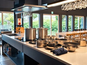 A modern kitchen with stainless steel appliances, a large island with cooking pots, and a view of greenery through large windows.