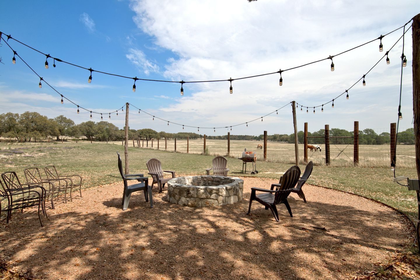 An outdoor seating area with chairs, a stone fire pit, and string lights against a backdrop of a fenced field and trees.