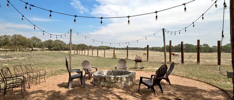 An outdoor seating area with chairs, a stone fire pit, and string lights against a backdrop of a fenced field and trees.