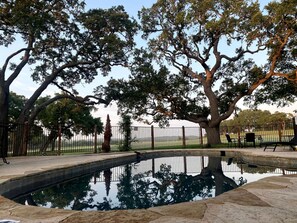A swimming pool surrounded by trees and a fenced area with outdoor seating.