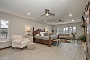 A bedroom with a large bed, two bedside tables, a chair, a bench, a ceiling fan, and a large window.
