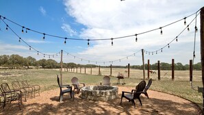 Un área de estar al aire libre con sillas, un hogar de piedra y luces colgantes, con un campo cercado y árboles de fondo.