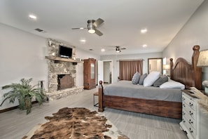 A bedroom with a stone fireplace, a bed with multiple pillows, a ceiling fan, and a wooden dresser. - Luxurious German-Style Ranch Vacation Rental with Pool Access and Stunning Views in Bergheim, Texas (Bergheim)