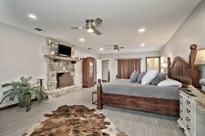 A bedroom with a stone fireplace, a bed with multiple pillows, a ceiling fan, and a wooden dresser. - Luxurious German-Style Ranch Vacation Rental with Pool Access and Stunning Views in Bergheim, Texas (Bergheim)