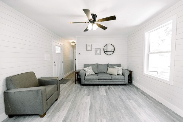 A room with a grey sofa, a grey armchair, a ceiling fan, and a large window.