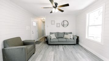 Una habitación con un sofá gris, un sillón gris, un ventilador de techo y un ventanal grande.