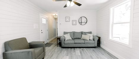 Una habitación con un sofá gris, un sillón gris, un ventilador de techo y un ventanal grande.