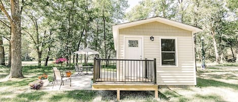 A small, single-story cabin with a front porch, surrounded by trees and a patio area with outdoor furniture.