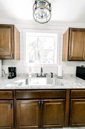 A kitchen with wooden cabinets, a stainless steel sink, a coffee maker, and a window with a view of trees.