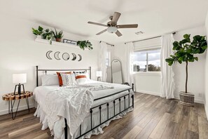 Un dormitorio con cama, mesitas de noche, ventilador de techo, ventana con cortinas y plantas en la pared.