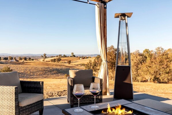 An outdoor seating area with wicker chairs, a table with glasses of red wine, a fire feature, and a tall, modern structure.