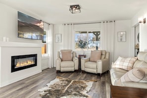 A modern living room with a fireplace, two armchairs, a sofa, a rug, and a large window with a view.