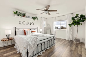 Un dormitorio con cama, mesitas de noche, ventilador de techo, ventana con cortinas y plantas en la pared.