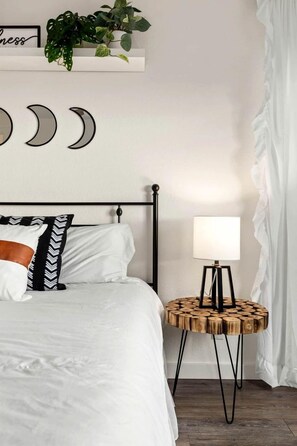 Un lit bien fait, avec une literie blanche, une tête de lit en métal noir et une table de chevet ronde en bois sur laquelle se trouve une lampe.