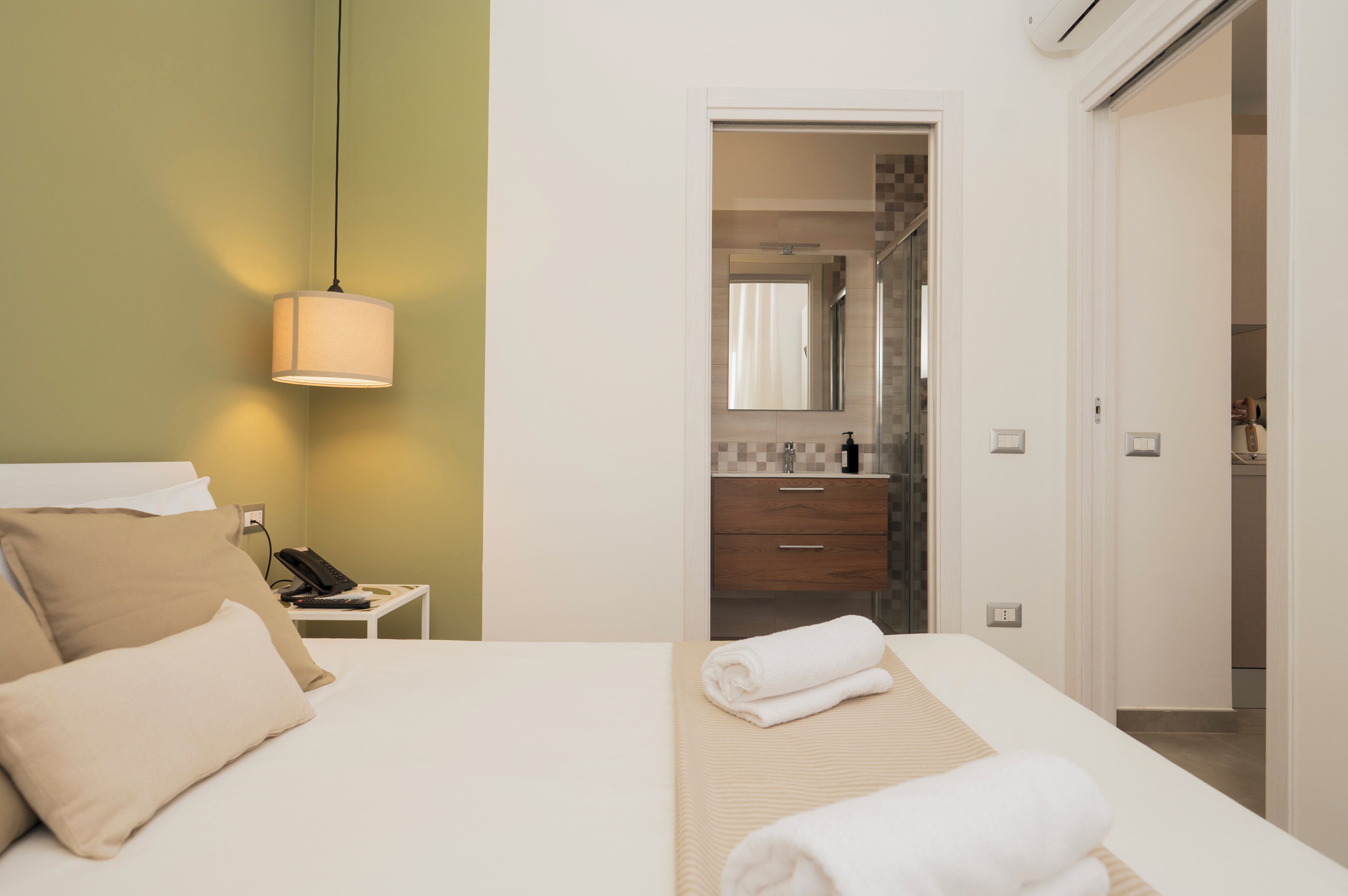 Camera d'albergo con un letto, un comodino con il telefono, una lampada e un bagno visibile attraverso una porta aperta.