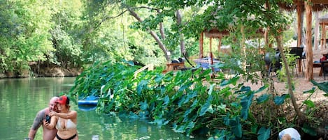 A couple sitting on a dock by a calm river, surrounded by lush greenery and a thatched-roof structure.