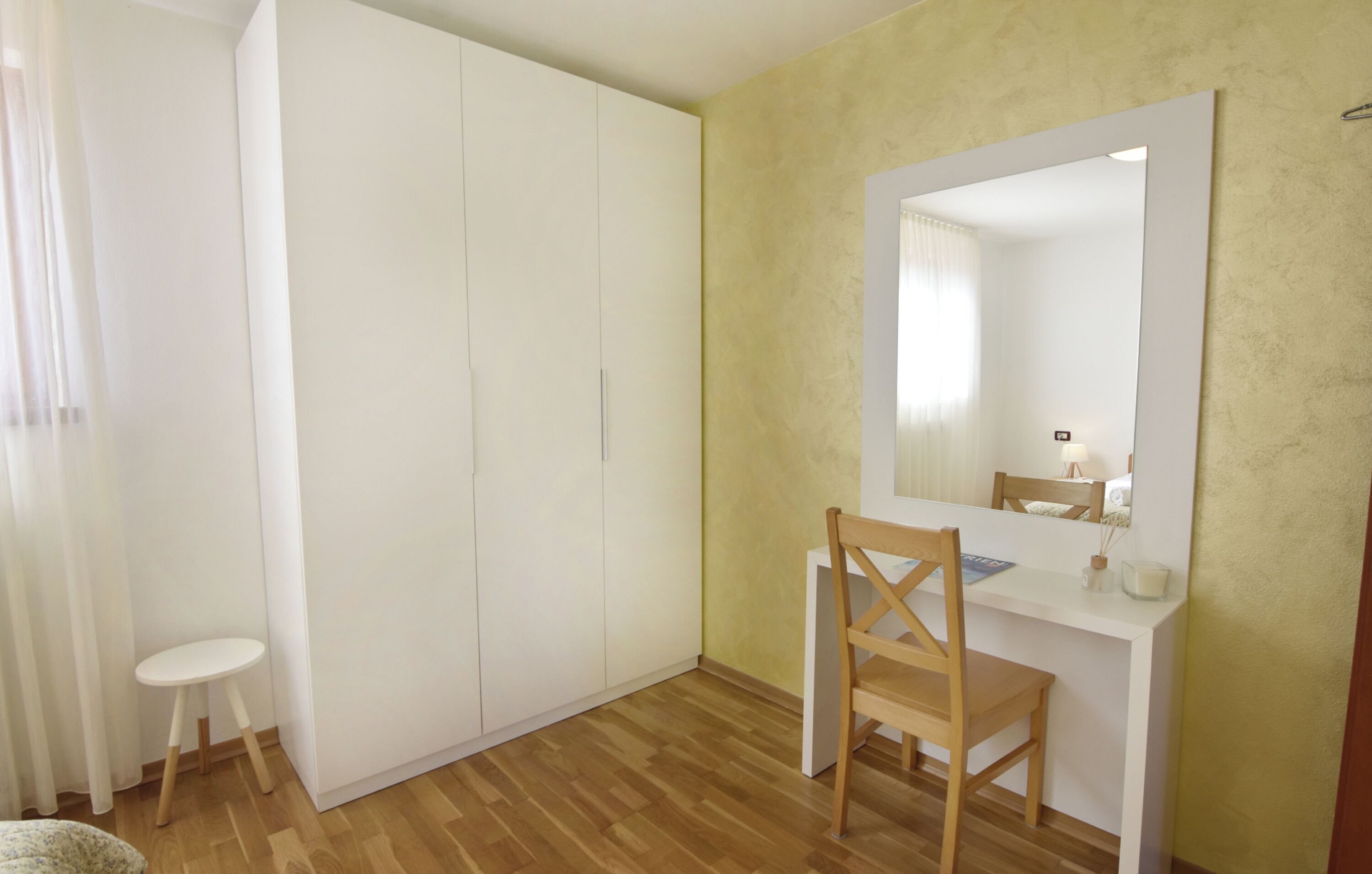 Et værelse med trægulv, et hvidt skrivebord med stol, en stor garderobe og et spejl på væggen.