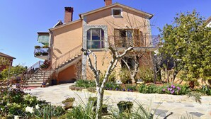 Una casa de dos pisos con fachada de piedra, una escalera principal y un jardín con diversas plantas y flores.