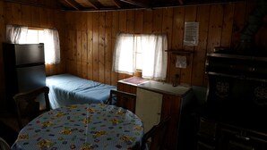 En liten stuga med en enkelsäng, ett litet kök, ett bord med en barnvänlig duk och ett fönster med gardiner.