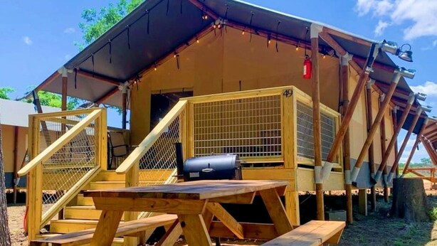 A wooden structure with a picnic table, a grill, and a fenced area. - Deluxe Safari Tent Cabin with Creek Swimming, Fishing & Resort-Style Amenities in Marion, Texas (Marion)