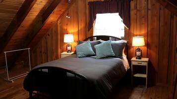 A cozy attic bedroom with a sloped ceiling, a bed with a headboard, two bedside tables with lamps, and a window with a curtain.