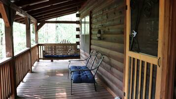 A wooden porch with a bench and a metal chair, a wooden door with a decorative glass panel, and a view of greenery outside.