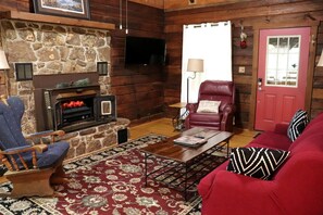 Ein Interieur einer Blockhütte mit Kamin aus Stein, einem an der Wand montierten Fernseher, einer roten Tür und einem roten Sofa mit gemusterten Kissen.