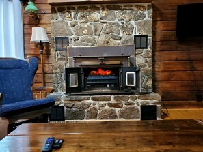 Ein Steinkamin mit Feuer, ein blau gepolsterter Sessel, ein Holztisch und eine Lampe.