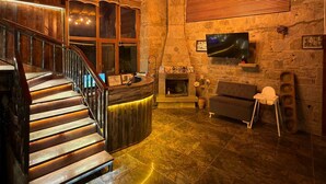 A wooden interior with a stone fireplace, a TV mounted on the wall, and a staircase with a wooden railing. - Assos Elit Pansiyon (Ayvacik)