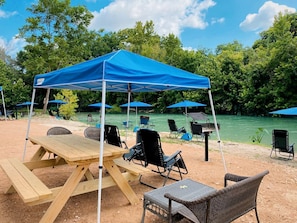 Ein Picknick am Strand mit Tisch, Stühlen, Sonnensegel und Blick auf einen Fluss.