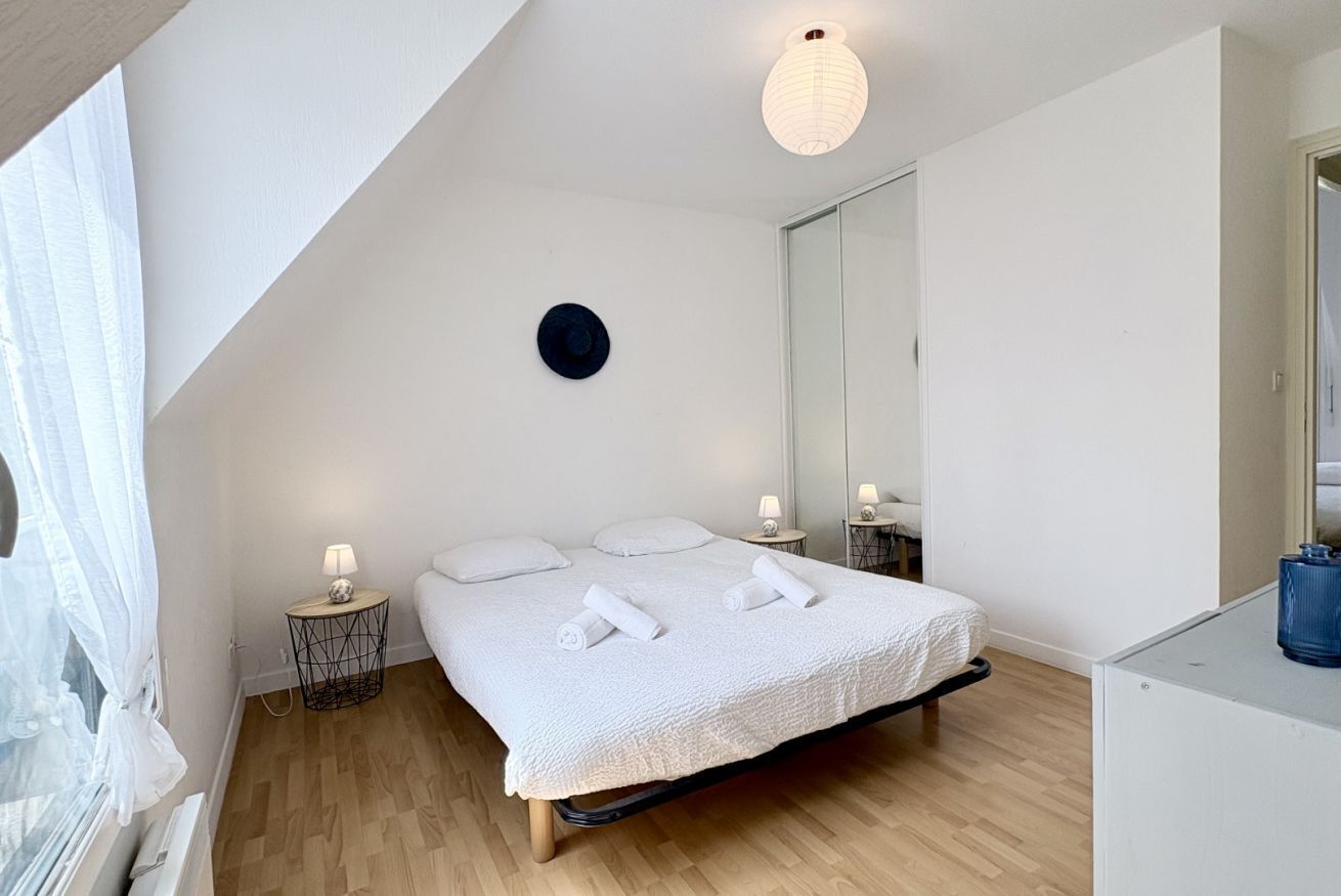 A bedroom with a bed, bedside lamps, a mirror, and a window with sheer curtains.