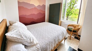 Ein Schlafzimmer mit Bett, Wandgemälde, Fenster mit Aussicht und einem Korbsessel.