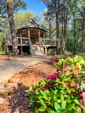 Eine Holzhütte mit überdachter Veranda und einer Treppe zu einer Terrasse, umgeben von Bäumen und einem Kiesweg.