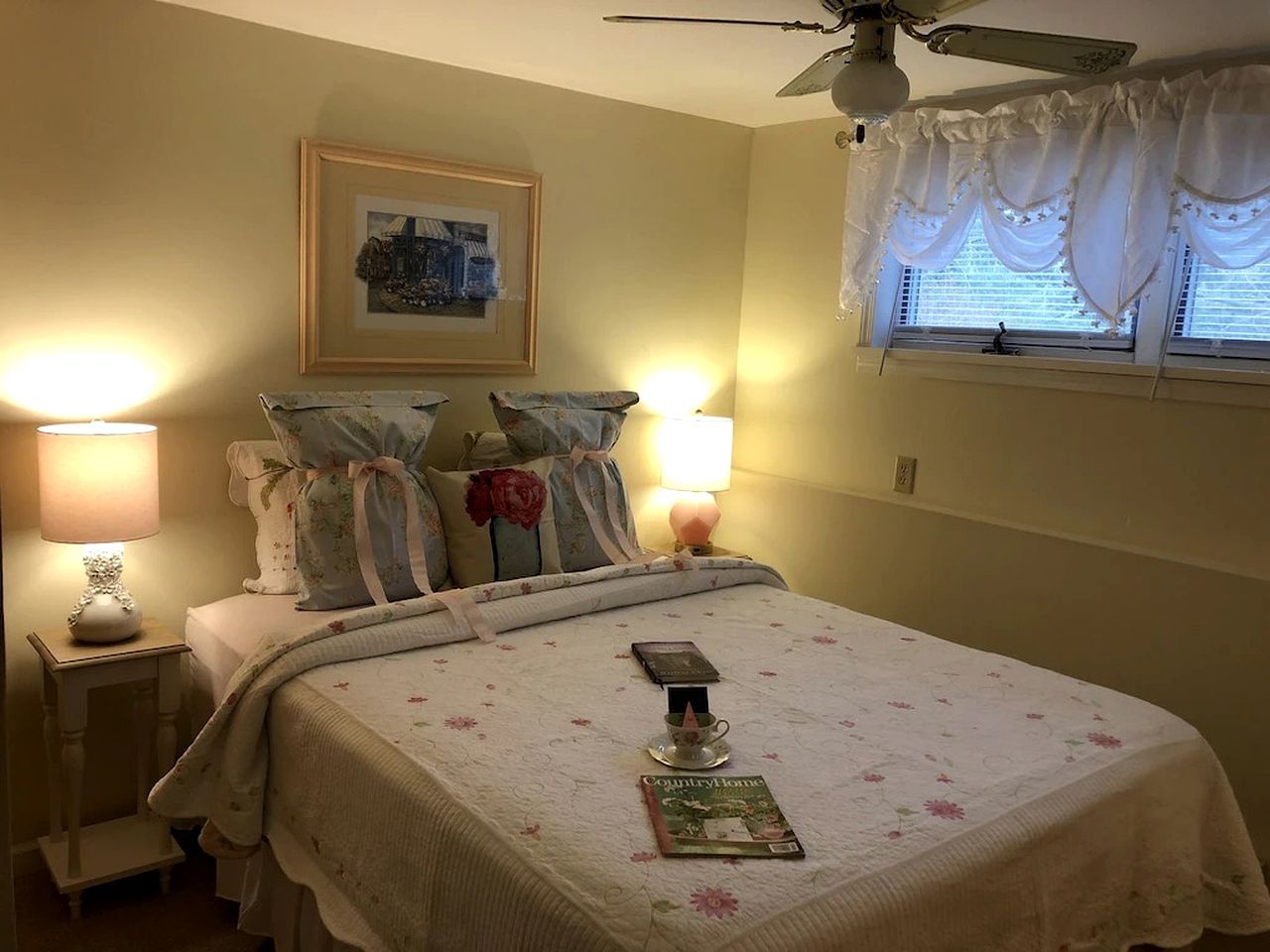 Un dormitorio con cama, lámparas de noche, un cuadro enmarco y una ventana con cortinas.