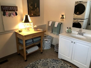A bathroom with a wooden vanity, a sink, a mirror, a towel rack, a laundry basket, and a wall-mounted laundry machine.
