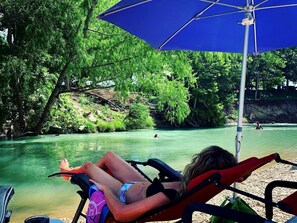 Eine Person entspannt sich in einem Liegestuhl unter einem blauen Sonnenschirm, im Hintergrund fließt ein Fluss und es gibt viel Grün.