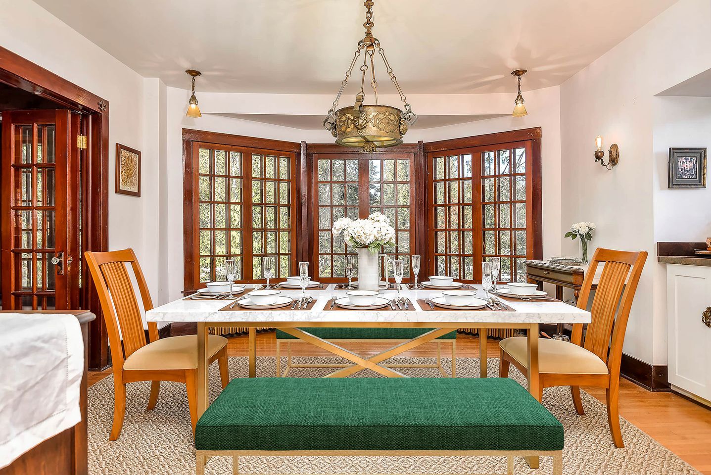 Une salle à manger avec une grande table dressée pour six personnes, entourée de chaises en bois, d’un banc vert et d’un lustre.