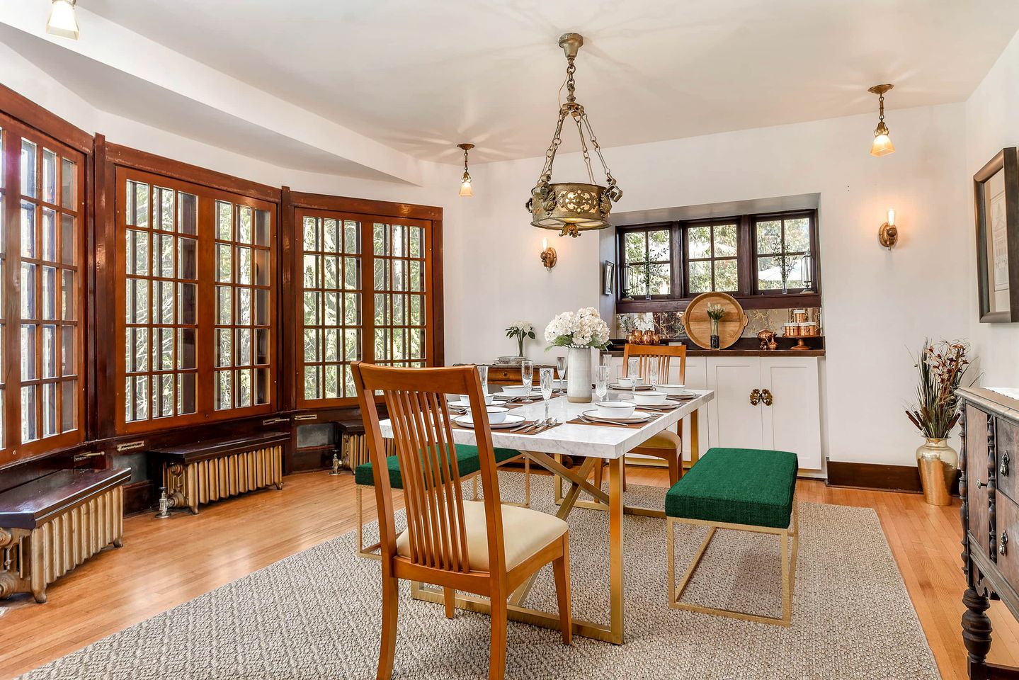 Un comedor con mesa de madera, sillas, una araña y ventanas grandes.