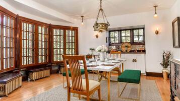 Un comedor con mesa de madera, sillas, una araña y ventanas grandes.