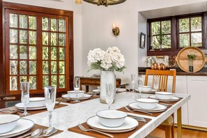 A dining room with a long table set for six, a vase of white flowers, and a view of the outdoors through large windows.